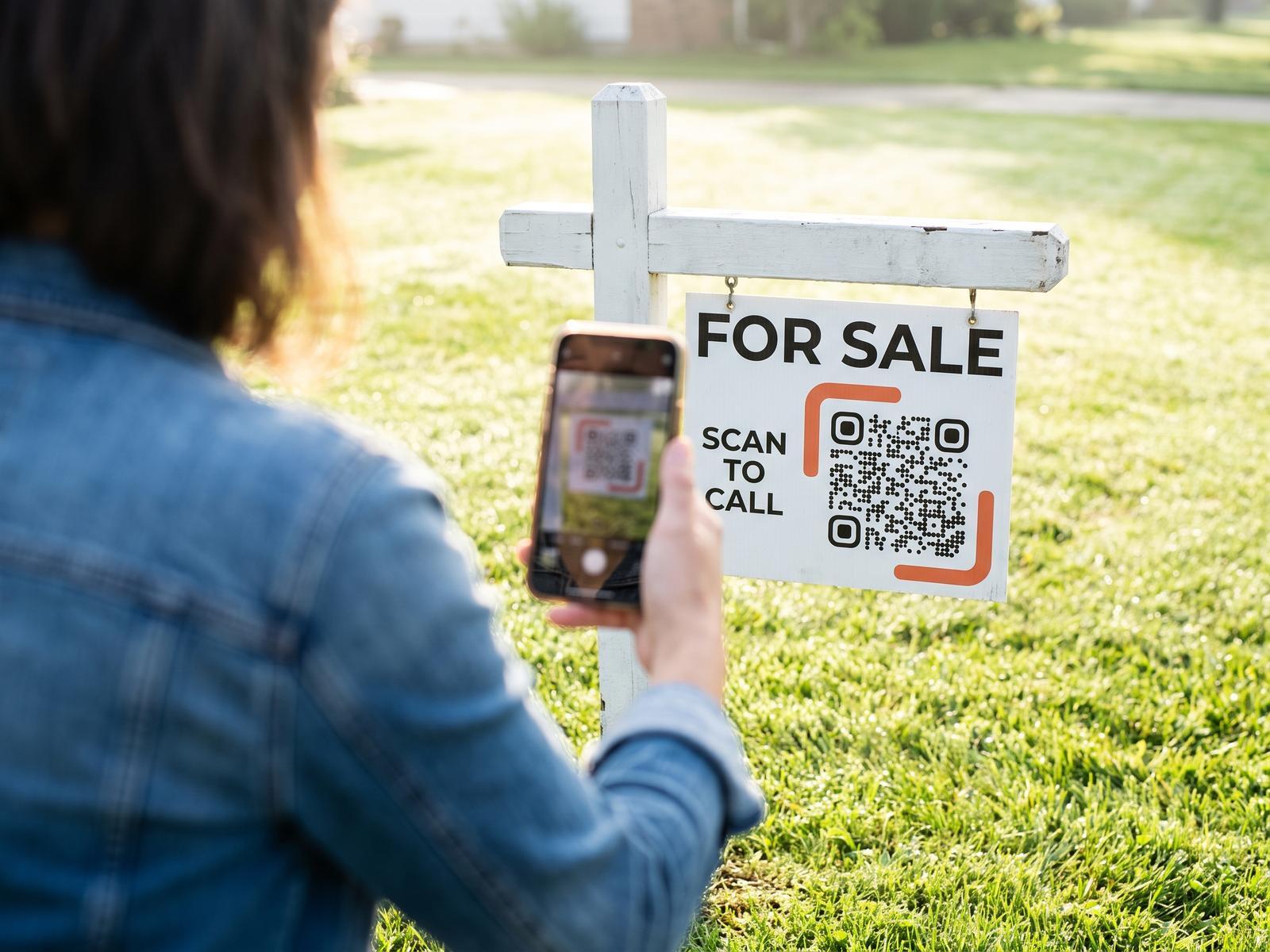 Someone scans the code on a “For sale · Scan to call” sign hanging from a post on a front lawn.