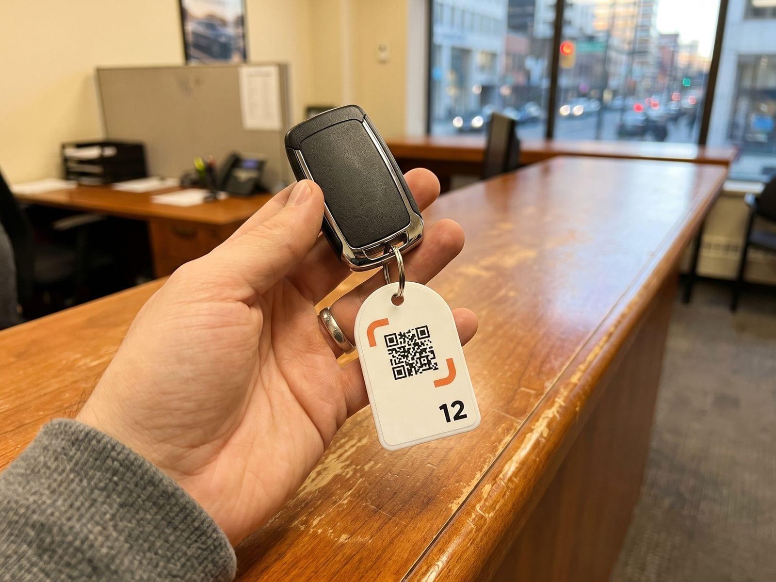 A hand holds a car key with a white key tag showing a QR code and the number 12.