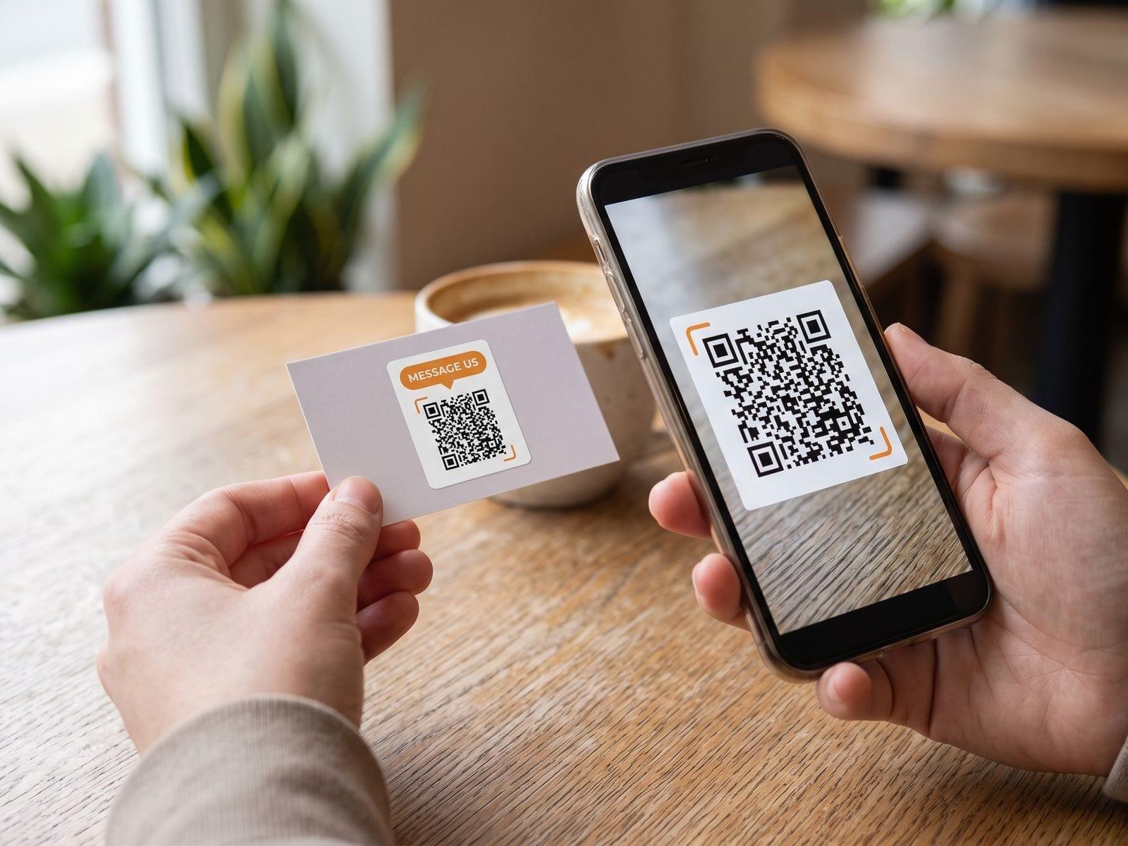 A “Message us” QR code sticker on the back of a business card, scanned with a phone across a café table.
