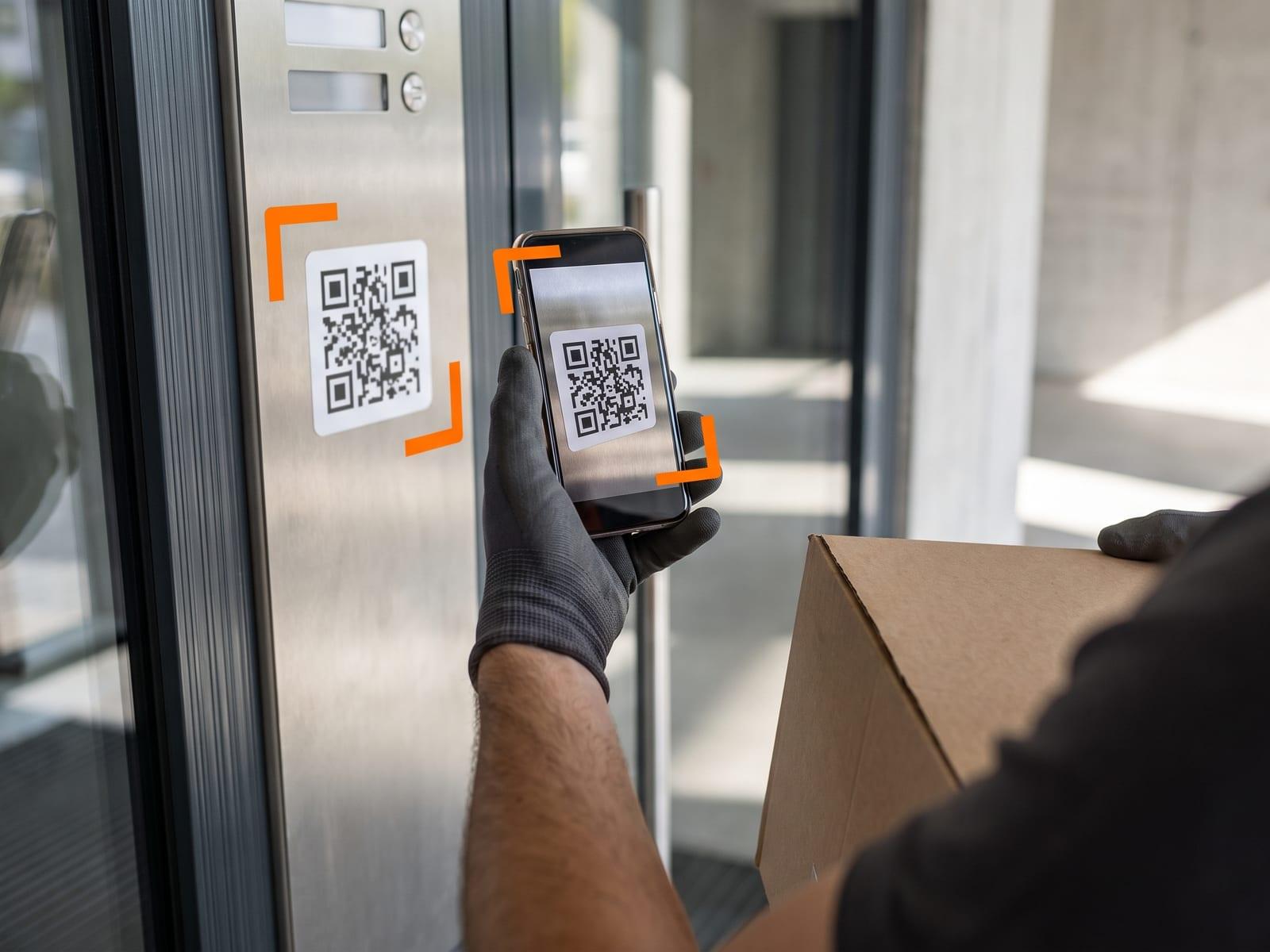 A courier’s gloved hand scanning a small doorbell sticker on a lobby intercom panel, a parcel under the arm.