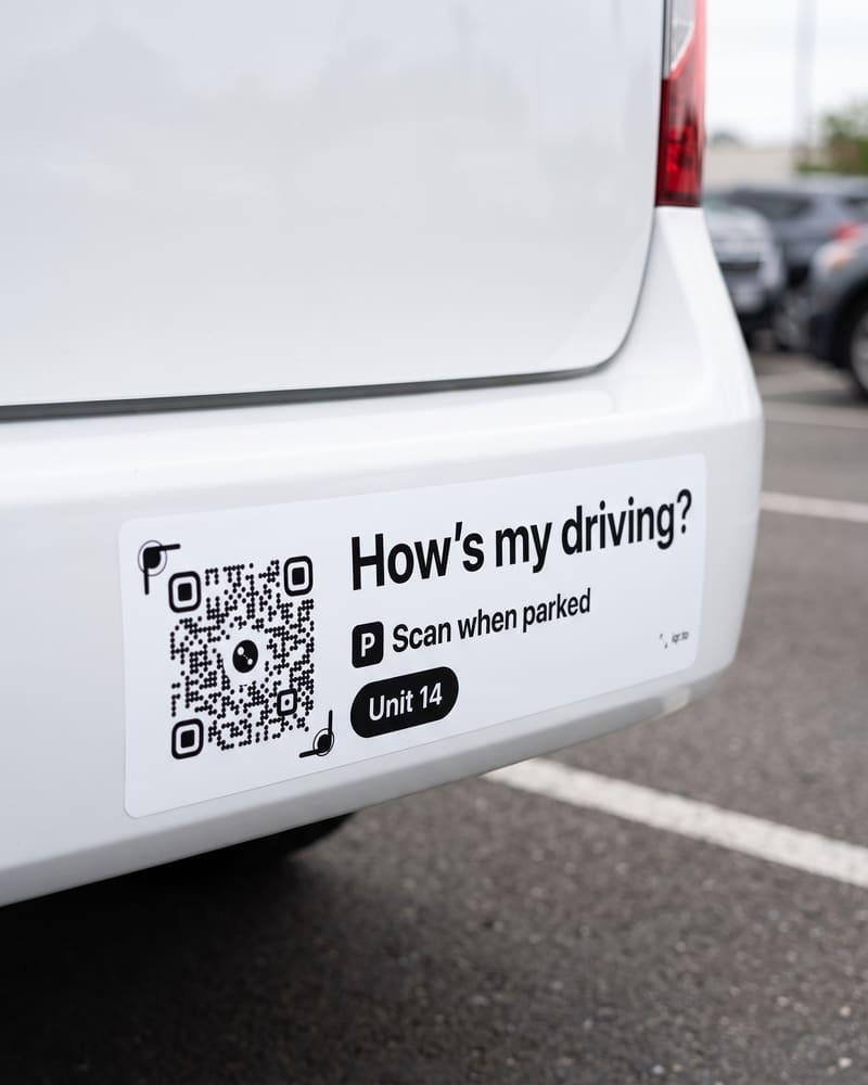 A long bumper sticker on a white van: How’s my driving, Scan when parked, Unit 14, and a QR code.