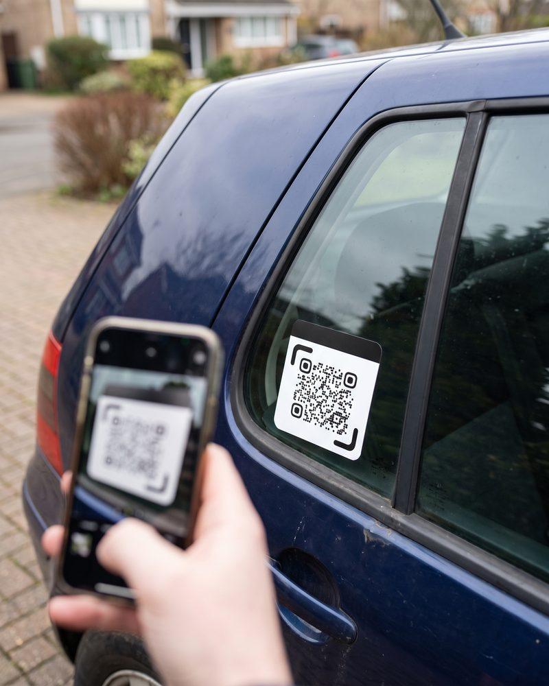 A hand holds a phone up to a small QR sticker on a car’s back window