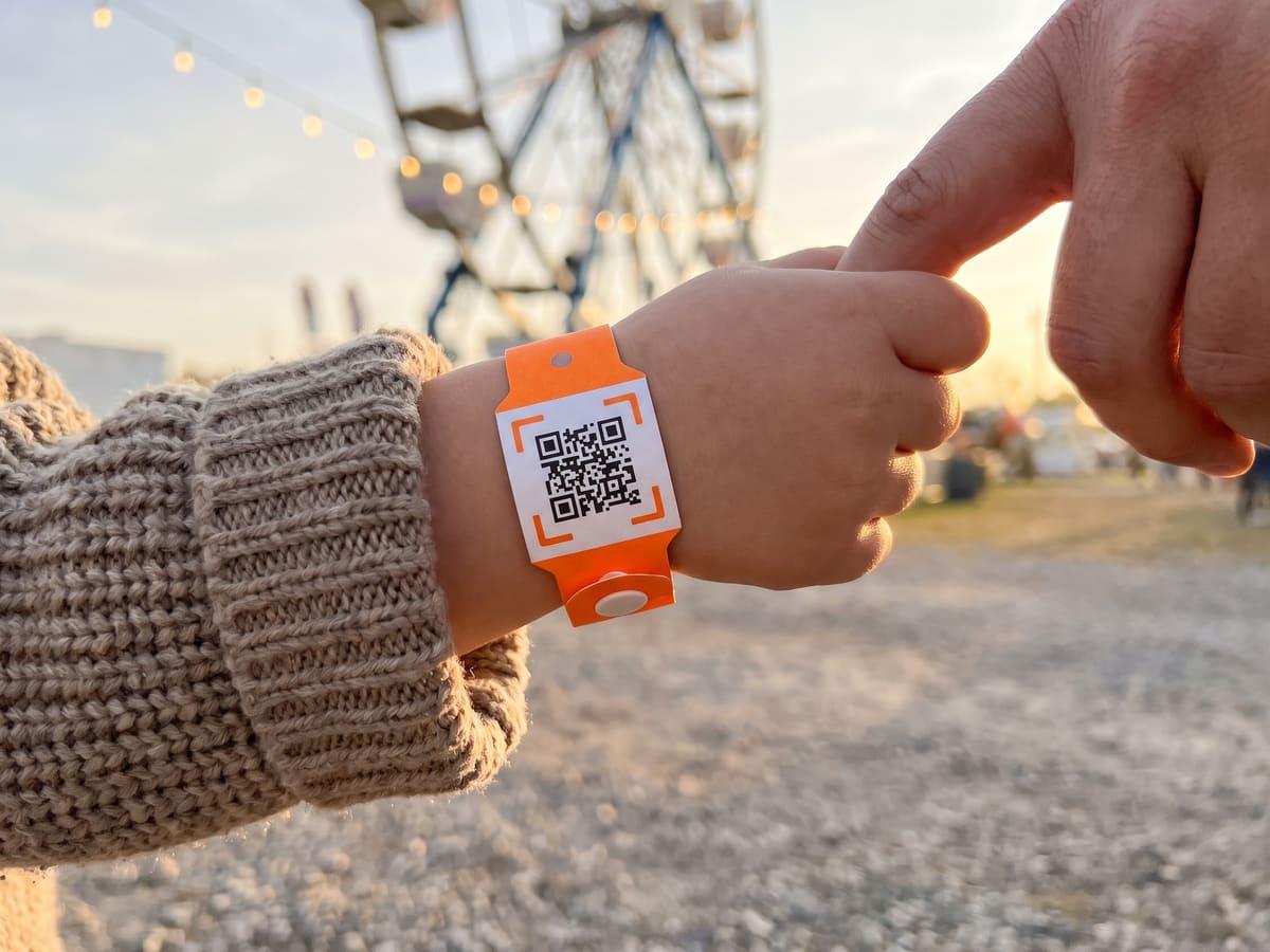 A child’s wrist with an orange paper wristband carrying an iQR code, holding a parent’s finger at a fair.