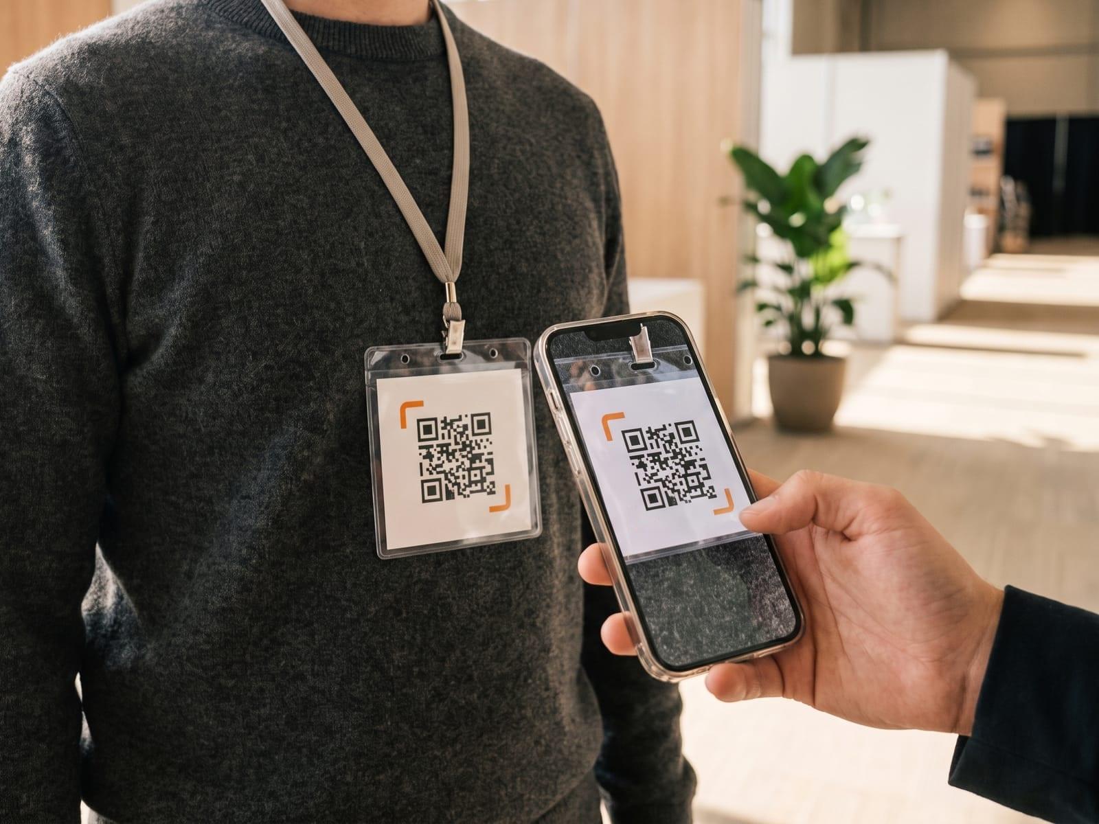 A conference badge with an iQR code sticker hangs from a lanyard; another person’s hand holds up a phone whose camera shows the code.