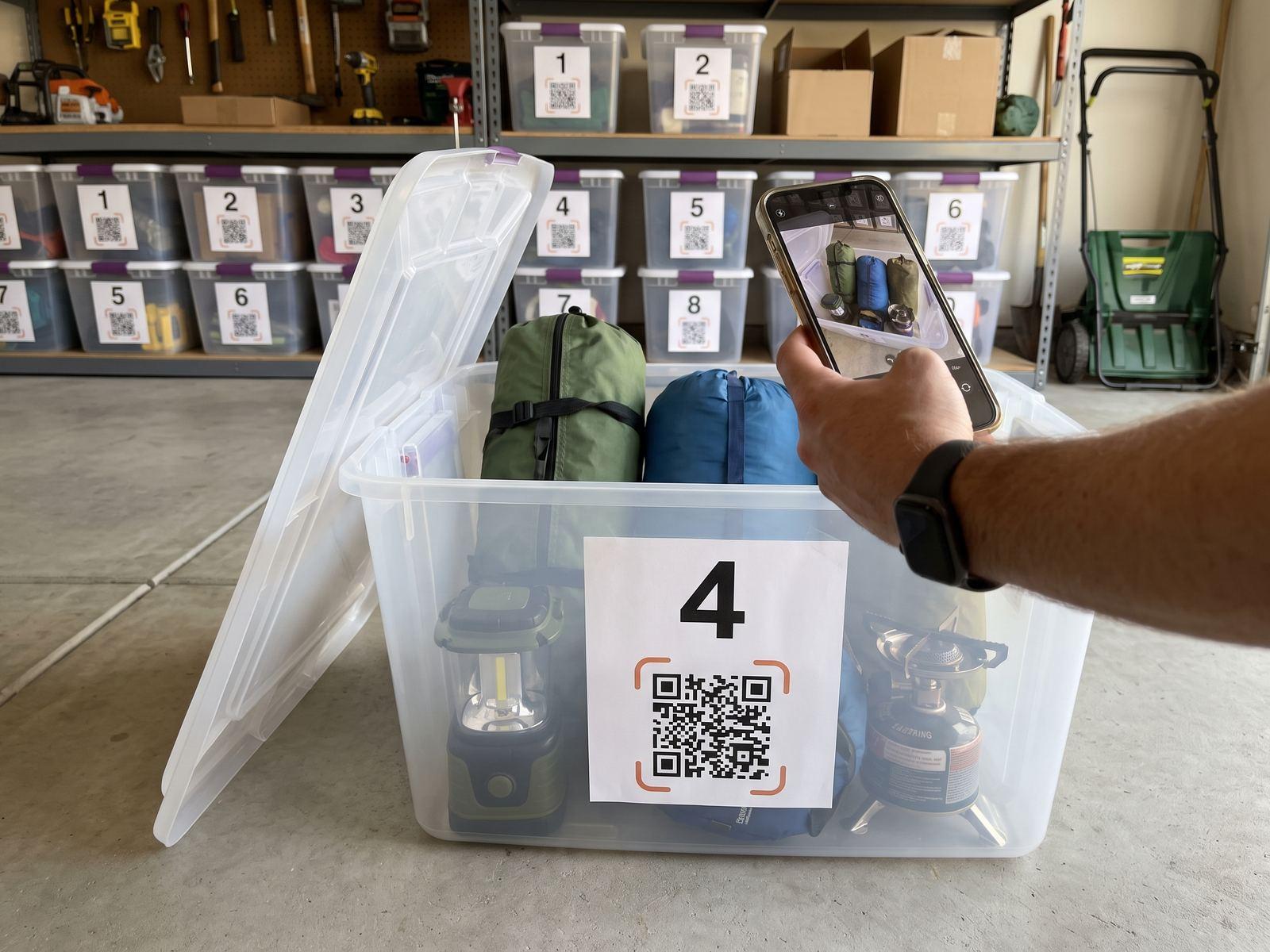 An open clear bin of camping gear on a garage floor, labelled 4 with a QR code, while a hand holds a phone over it to photograph what is inside.