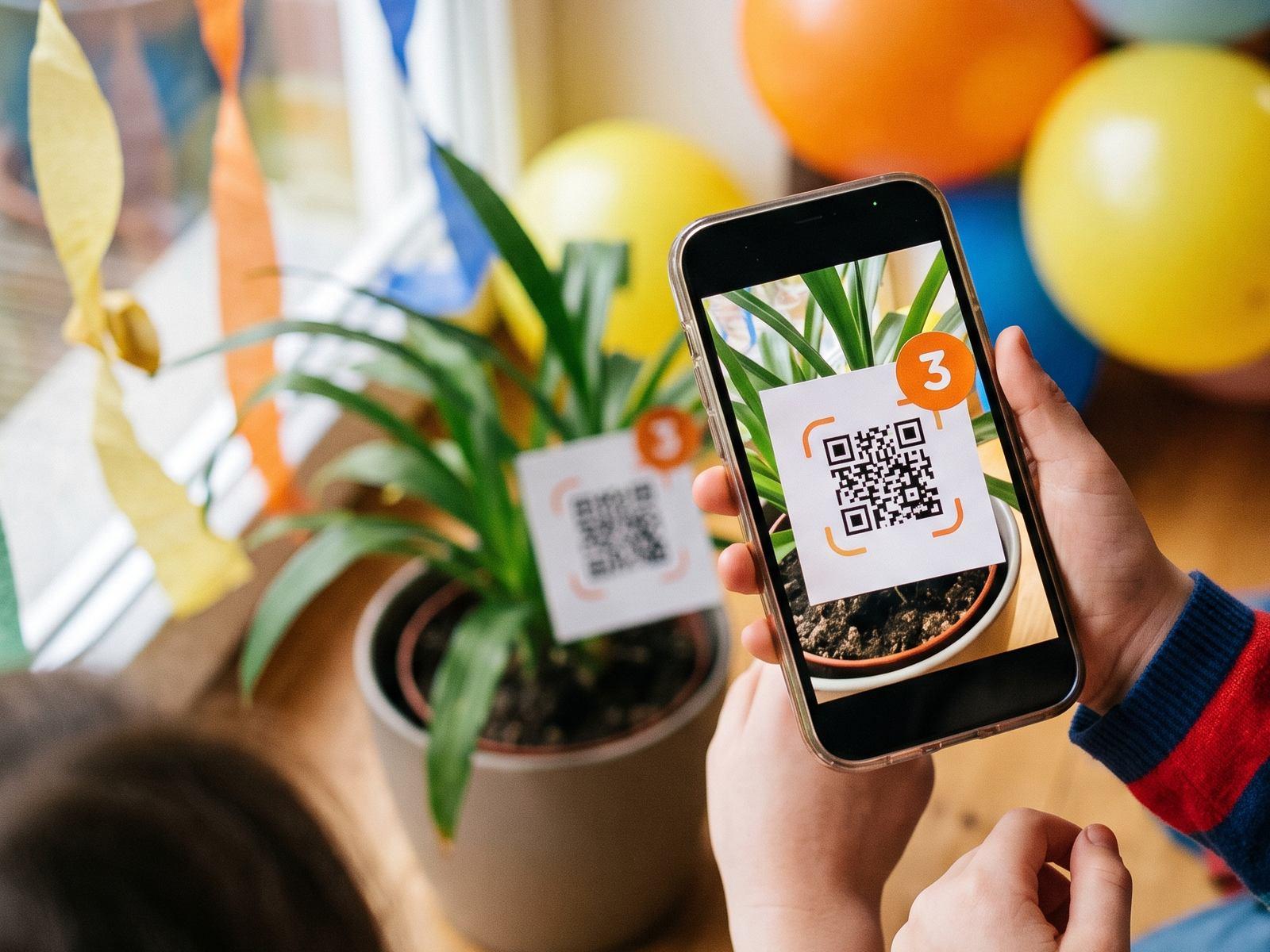At a birthday party, a child’s phone camera frames checkpoint 3, a QR code card tucked into a plant pot