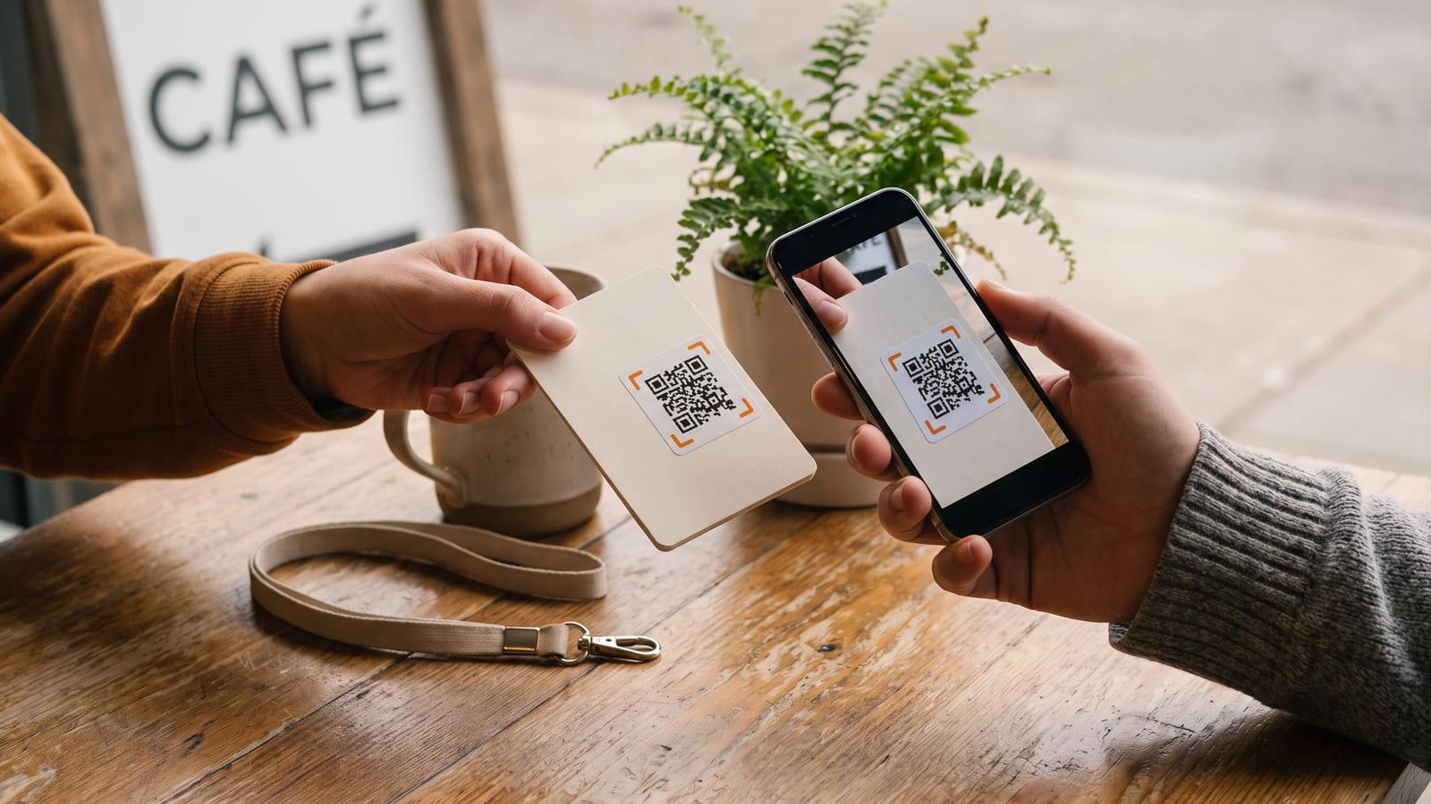 One person handing a business card with a QR code across a café table while the other scans it, a lanyard beside the coffee.