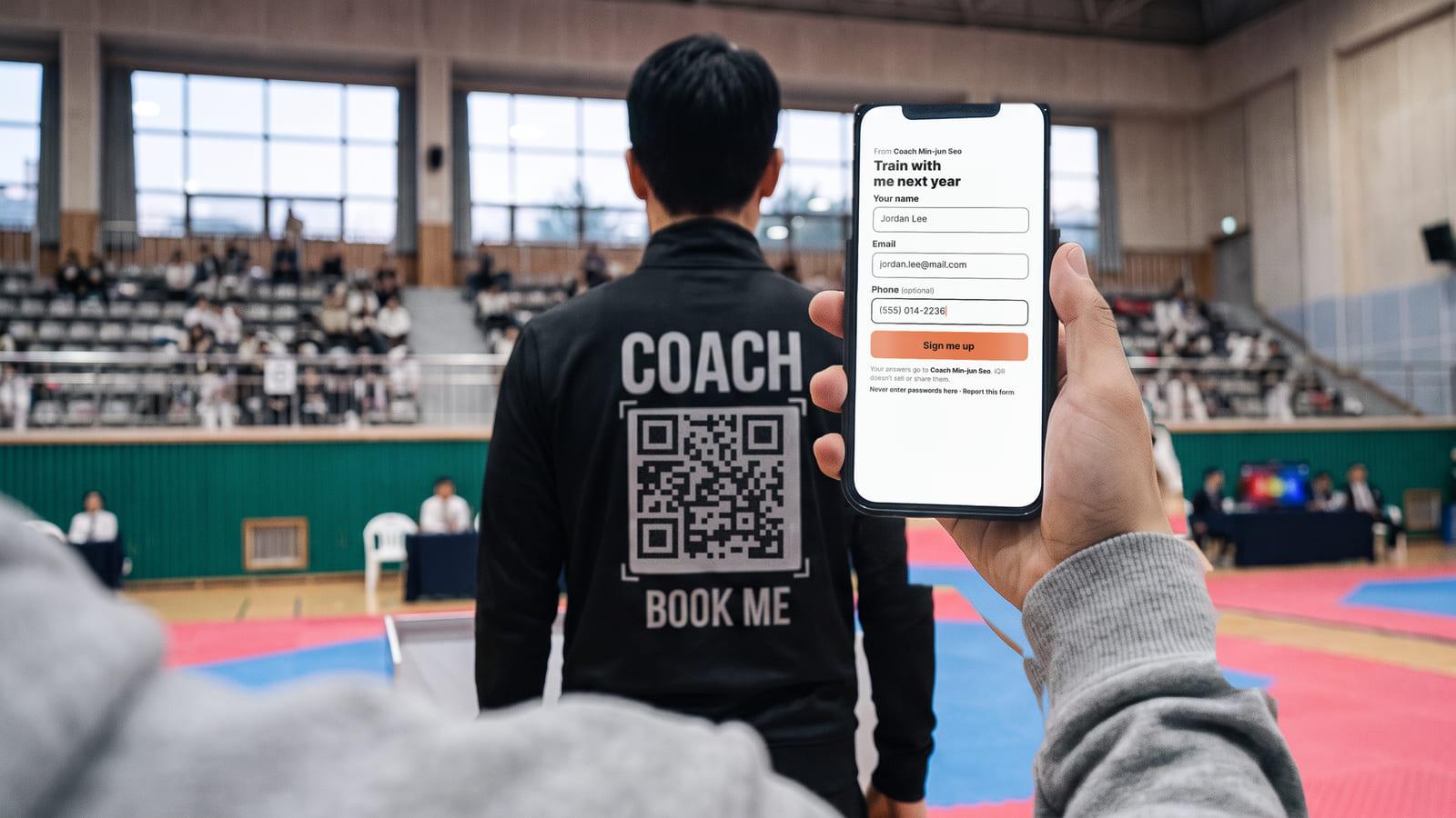 A taekwondo coach seen from behind at the edge of the mat, his black track jacket printed COACH, a QR code and BOOK ME, while a spectator holds up a phone showing his sign-up form filled in.