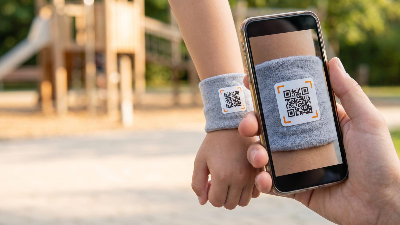 A child safety wristband with a QR code on a child’s wrist at a playground, an adult’s phone scanning it.